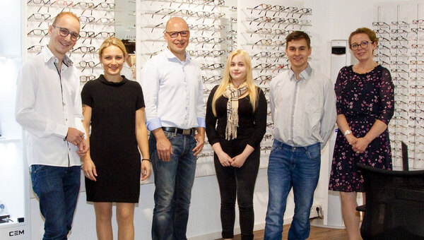 Sechs Personen stehen in einem modernen Optikerladen vor einer Wand mit vielen Brillen.